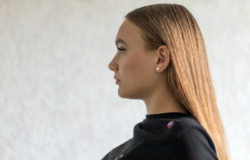 A woman with straight, light brown hair and a black top is shown in profile against a light, plain background.