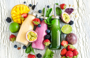 Three glass bottles of smoothies—yellow, purple, and green—surrounded by assorted fresh fruits including mango, strawberries, lime, and passion fruit on a white wooden surface.