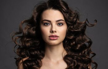 A woman with long, voluminous dark brown curly hair looks directly at the camera against a plain dark background.