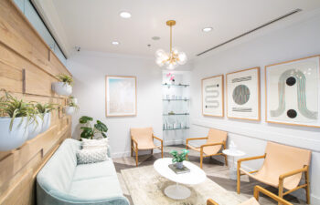 A modern waiting room with a light sofa, tan chairs, a round white coffee table, wall art, potted plants, and soft overhead lighting.