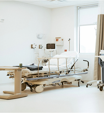A hospital room with an empty bed, medical equipment, and a side table. The room is well-lit with a window and has a neutral color scheme.