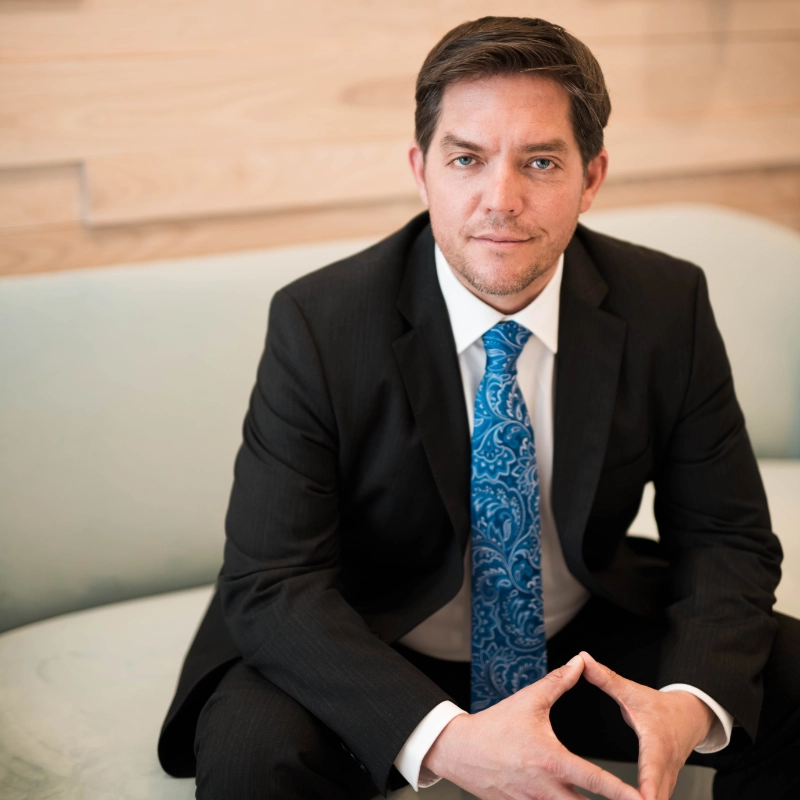 Dr. Benjamin C. Stong in a suit with a blue tie sitting on a light-colored sofa, hands clasped, in front of a wooden background.