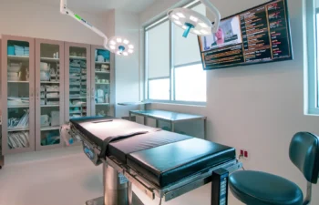 Interior of an empty surgical operating room with a black operating table, cabinets stocked with medical supplies, overhead lights, and a wall-mounted screen displaying information.