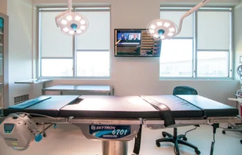 Medical operating room with an empty surgical table, overhead lights, a monitor displaying software, and large windows in the background.