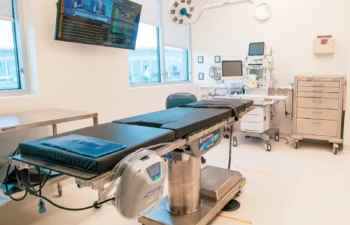 Hospital operating room with a surgical table, overhead light, medical monitors, and equipment carts. A TV screen displays news.