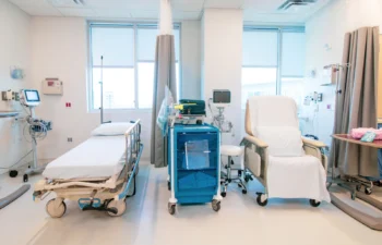 Hospital room with a bed, medical equipment, a blue cart, and a reclining chair. Curtains partially divide the space. Bright lighting from large windows.