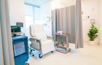 A hospital room with a reclining chair, medical equipment, a small table, and a privacy curtain. A potted plant is in the corner by the window.