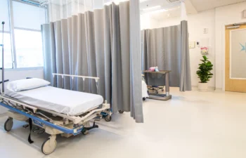 Hospital room with an empty bed, IV pole, tray table, and plant. The space is divided by grey privacy curtains, with a closed door and exit sign visible in the background.