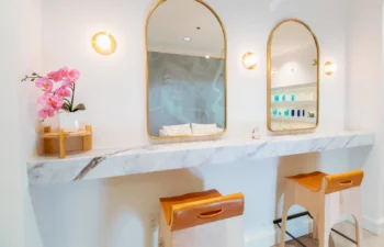 Two wooden stools sit at a marble counter with two oval mirrors and soft lighting. A pink orchid decorates the left side.