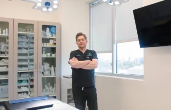 A person stands with arms crossed in a medical office, featuring cabinets of supplies, ceiling lights, a window, and a wall-mounted TV.