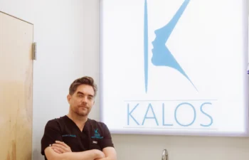 A person in medical scrubs stands with arms crossed in front of a sign displaying a stylized face profile and the word "Kalos" in an office setting.
