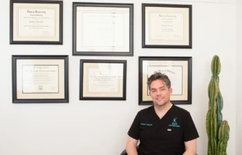 A man in a black scrub top sits in an office with framed certificates on the wall behind him, next to a tall cactus.