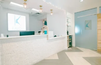 Bright reception area with a modern white counter, hanging lights, and decorative wall. A door with a logo and a visible sign reading "Kalos" are in the background.