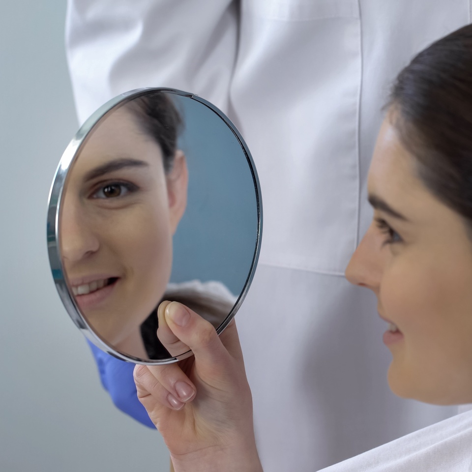 Young female satisfied with rhinoplasty result, smiling face reflected in mirror