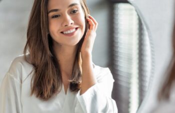Beauty Concept. Portrait Of Attractive Happy Woman Looking At Mirror In Bathroom, Beautiful Millennial Lady Wearing White Silk Robe Smiling To Reflection, Enjoying Her Appearance, Selective Focus