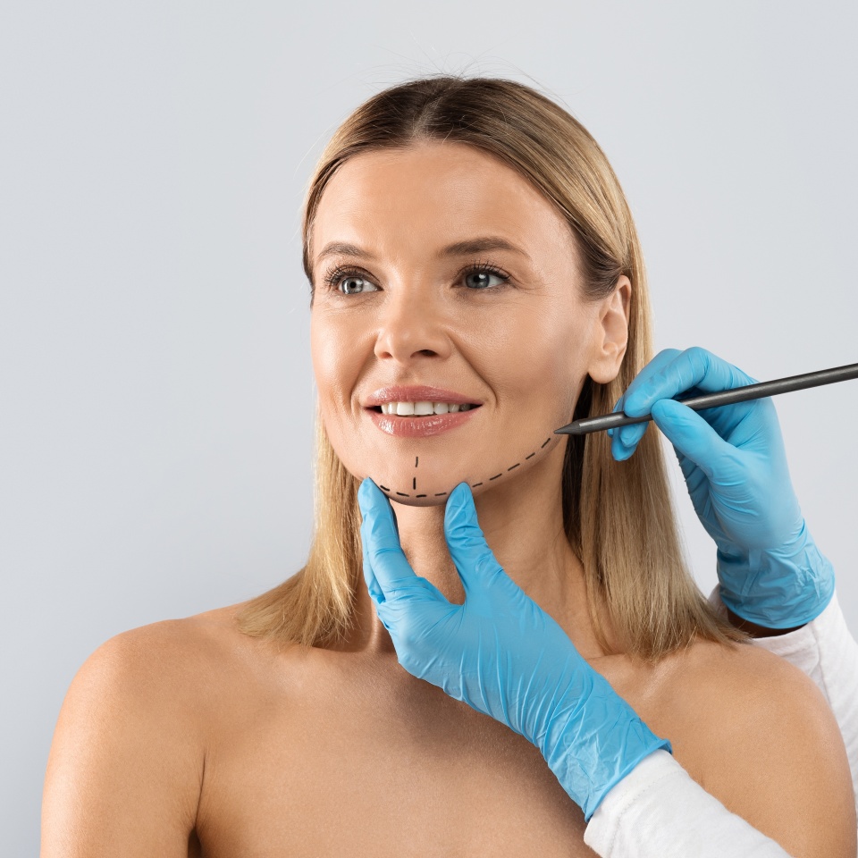 Hands of plastic surgeon wearing blue medical gloves drawing contours with black pen around female chin before surgery, middle aged blonde woman getting face-lift plastic surgery, isolated on grey