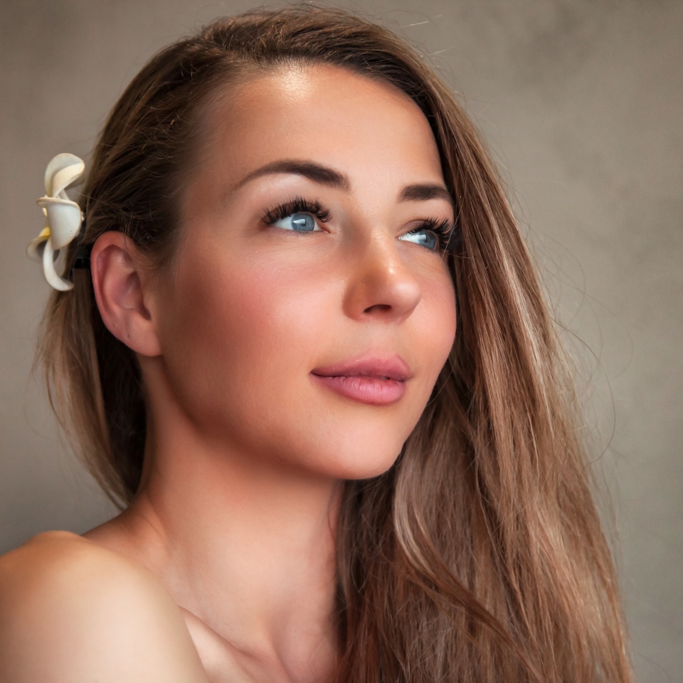 Soft light gently highlights a woman with long, flowing hair and a delicate flower tucked behind her ear. She gazes thoughtfully into the distance, radiating calm in an inviting indoor space.