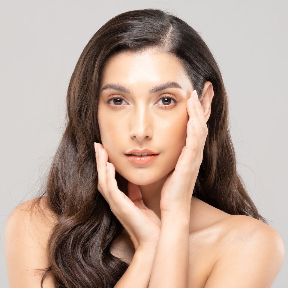 A woman with long brown hair poses against a neutral background, resting her hands gently on her cheeks, and looking directly forward.