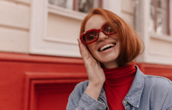 A woman with red hair wearing red sunglasses, a red turtleneck, and a denim jacket smiles while touching her face, standing in front of a red and cream building.