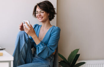 A person with short brown hair and glasses, wearing a blue top and jeans, sits by a desk and smiles while looking at a smartphone. A green plant is beside them.