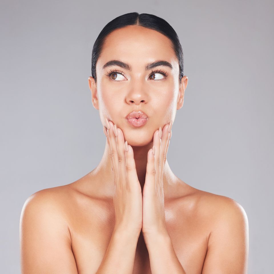 Person with a slicked-back hairstyle, making a playful expression while touching their face with both hands against a gray background.