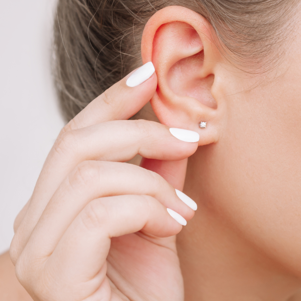 A close-up of a person’s ear wearing a small stud earring. The hand with white polished nails is gently touching the earlobe.