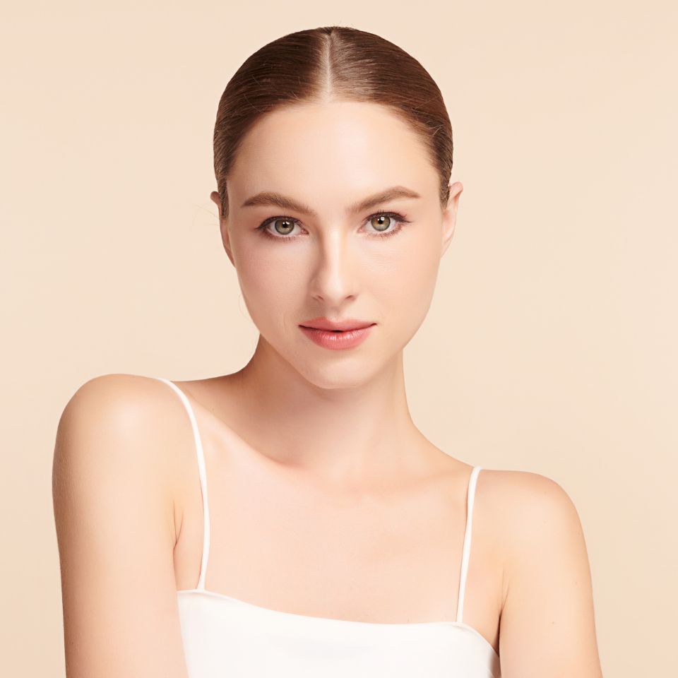 A woman with brown hair in a slicked-back style wears a white tank top and looks directly at the camera against a beige background.