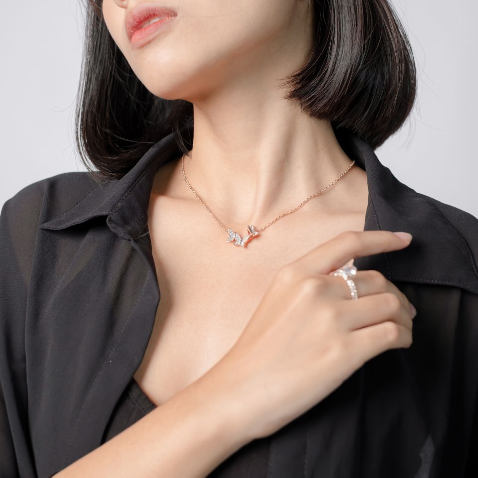 Person wearing a black blouse and a gold necklace with a leaf design, their right hand touching their neck and showcasing a ring with a large, round clear gemstone.