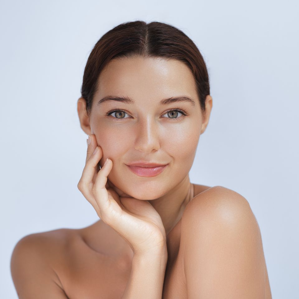 A woman with dark hair poses against a light background, resting her chin on her hand. She has a neutral expression and bare shoulders.