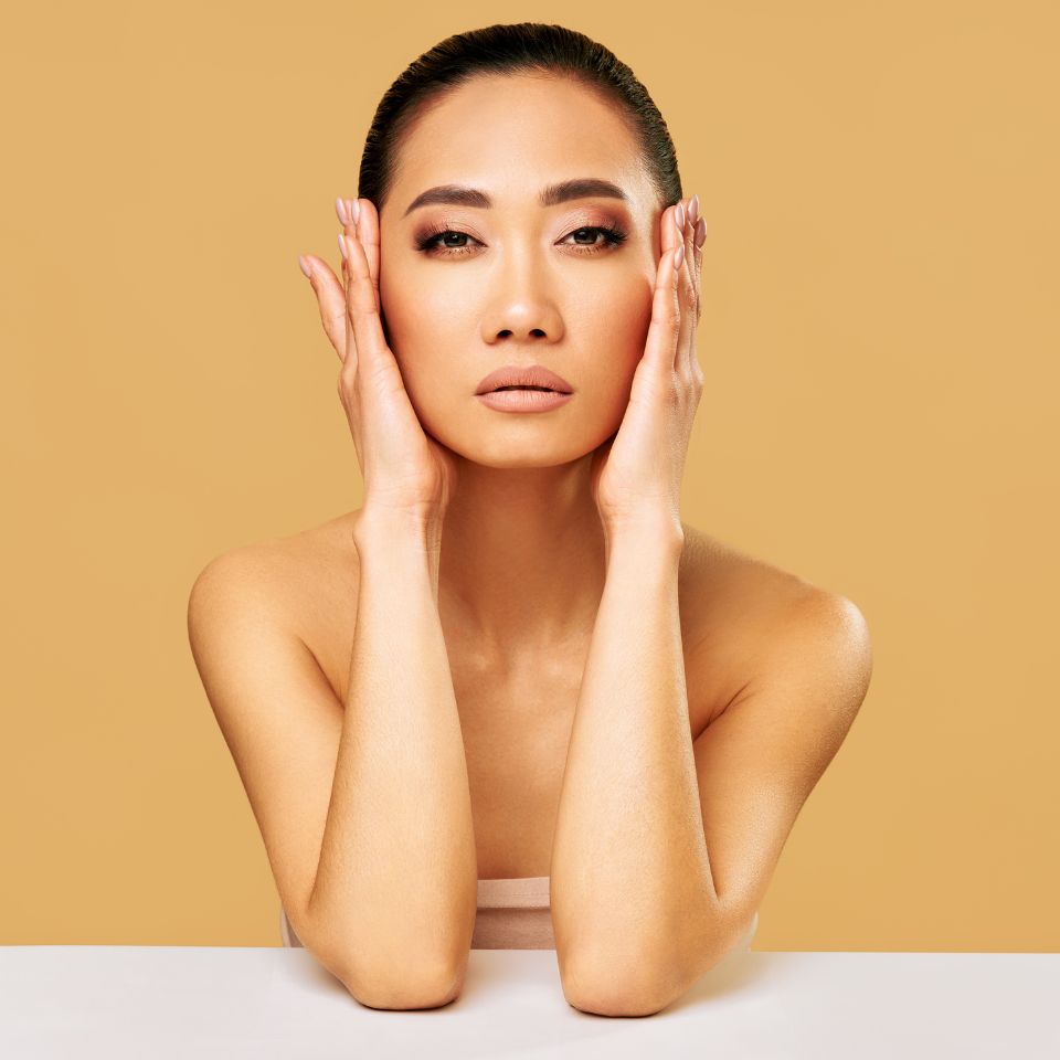 A person with long hair pulled back poses against a beige background, resting hands on face, looking directly at the camera.