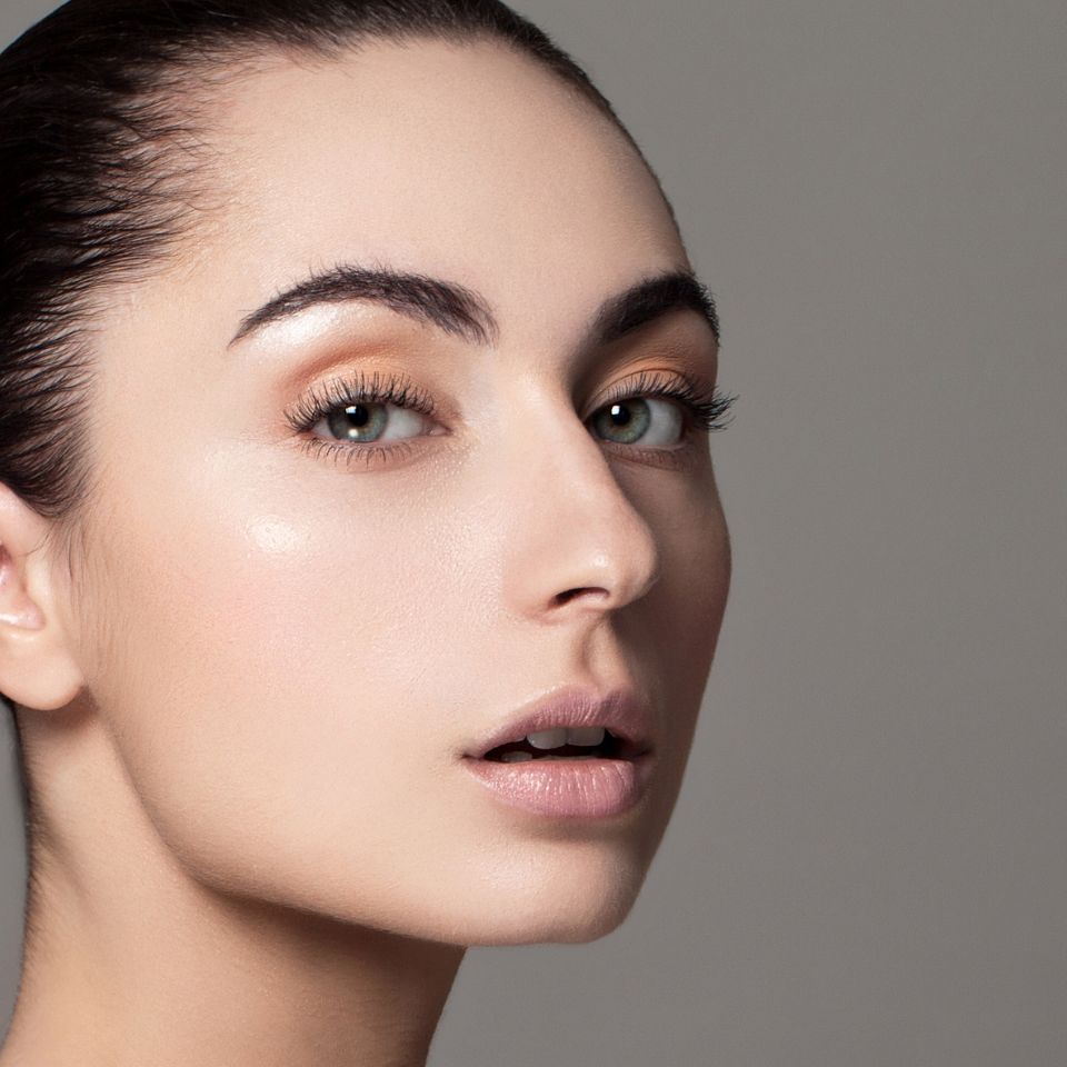 A close-up of a person with light makeup, soft eyebrows, and glossy lips, looking at the camera against a gray background.