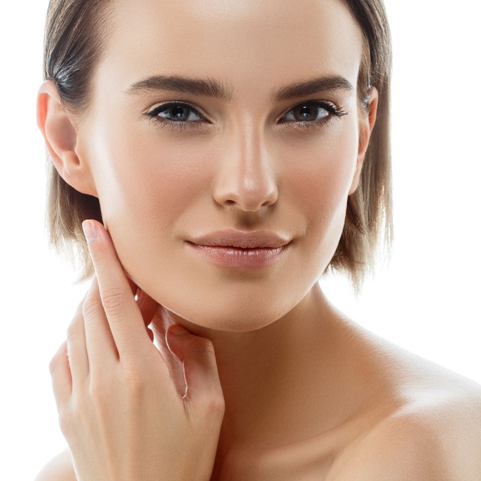 A woman with short hair gently touches her face, looking directly at the camera with a neutral expression against a white background.