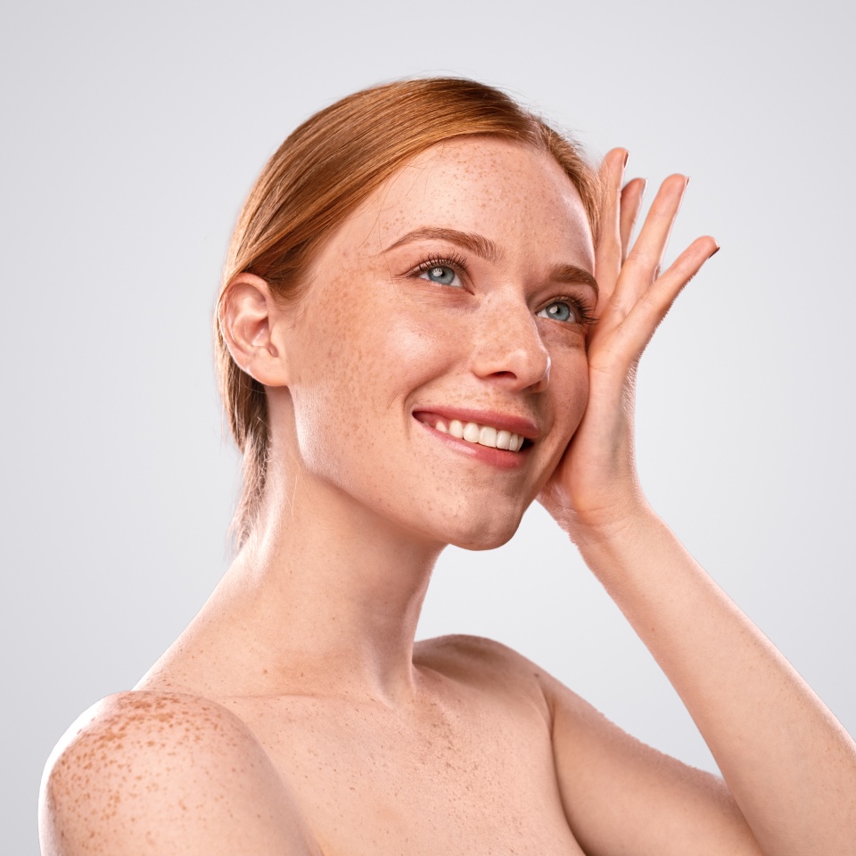 Smiling young ginger female with bare shoulders touching face and enjoying clean healthy skin on gray background
