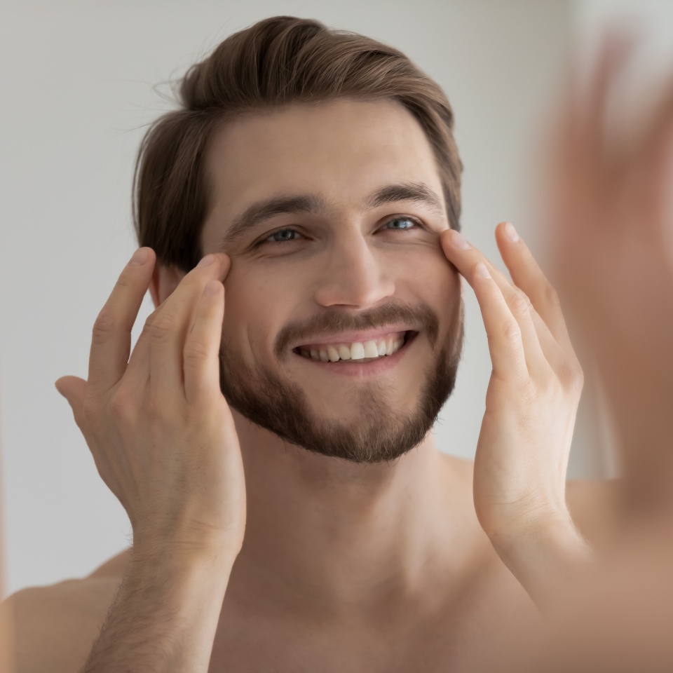 Close up mirror reflection smiling satisfied young man touching skin under eyes, applying anti aging moisturizing cream or lotion, standing in bathroom, enjoying skincare procedure, morning routine