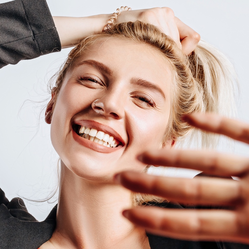 A beautiful blonde girl holds her hair and makes her own hairstyle with the emotion of happiness. Woman pulls her hand towards the camera on a gray isolated background for advertising. Nose piercing.