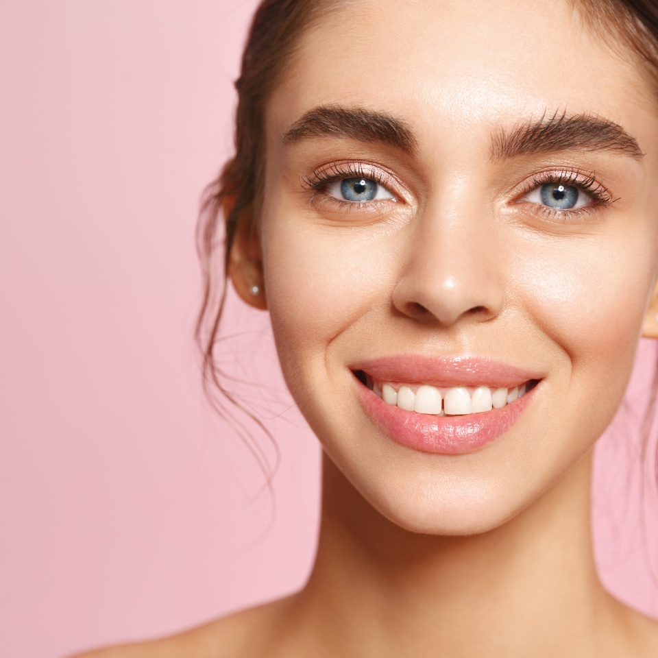 Portrait of caucasian young woman with blue eyes and perfect white smile, nude lips gloss and no makeup natural look, gazing happy at camera with clean hydrated facial skin, pink background.