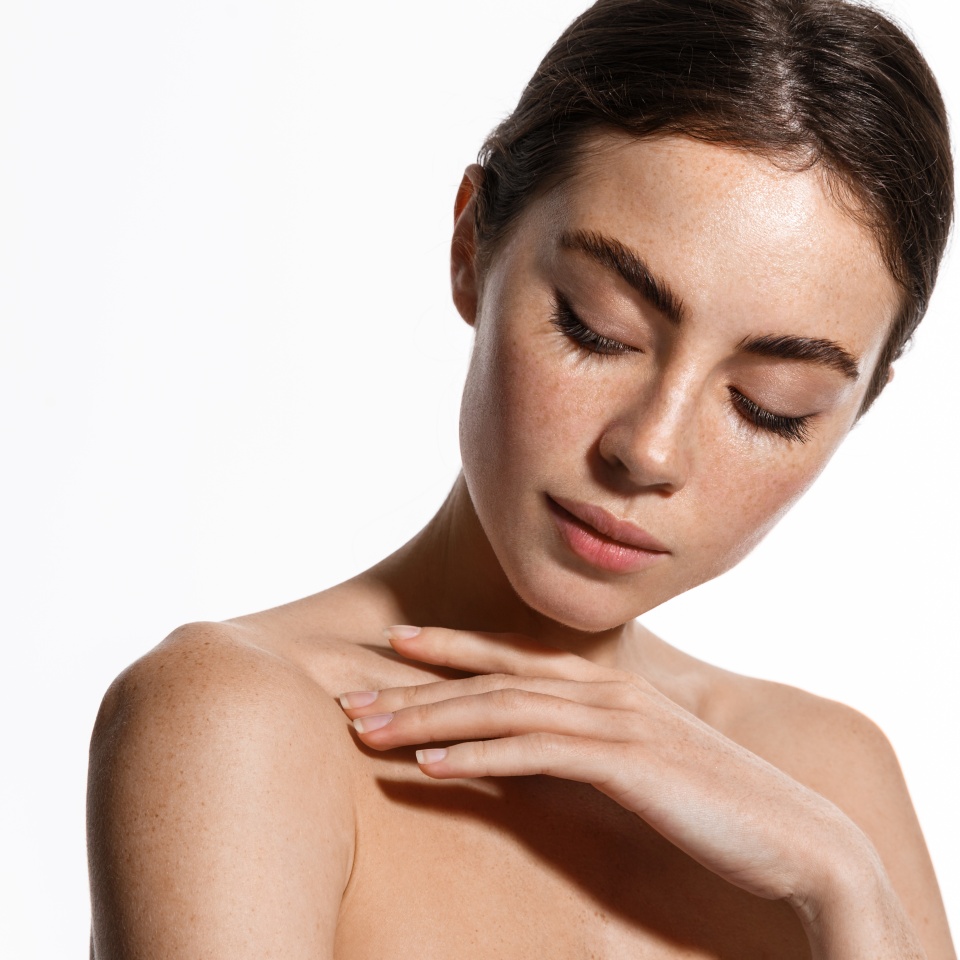 Young woman applying soft skin care cream on her body after shower, gently touching shoulder with fingertips, standing against white background, perfect healthy body and skin