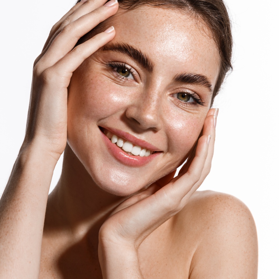 Vertical portrait of beautiful girl smiling at camera, touching natural face without makeup, healthy glowing facial skin and white teeth, standing over white background
