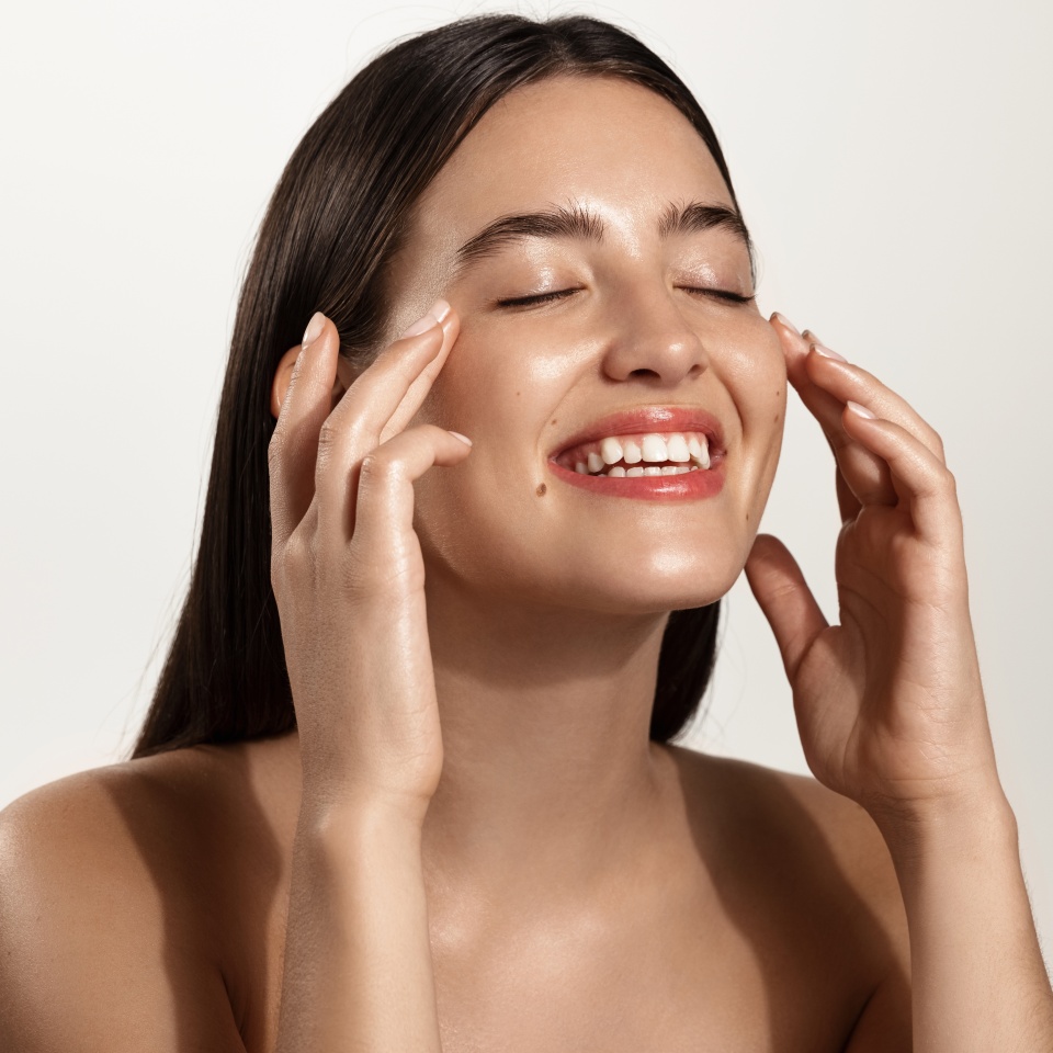 Smiling woman with clean glowing facial skin washing her face, rubbing in cream skincare product for anti-aging, lifting spa effect, standing over white background