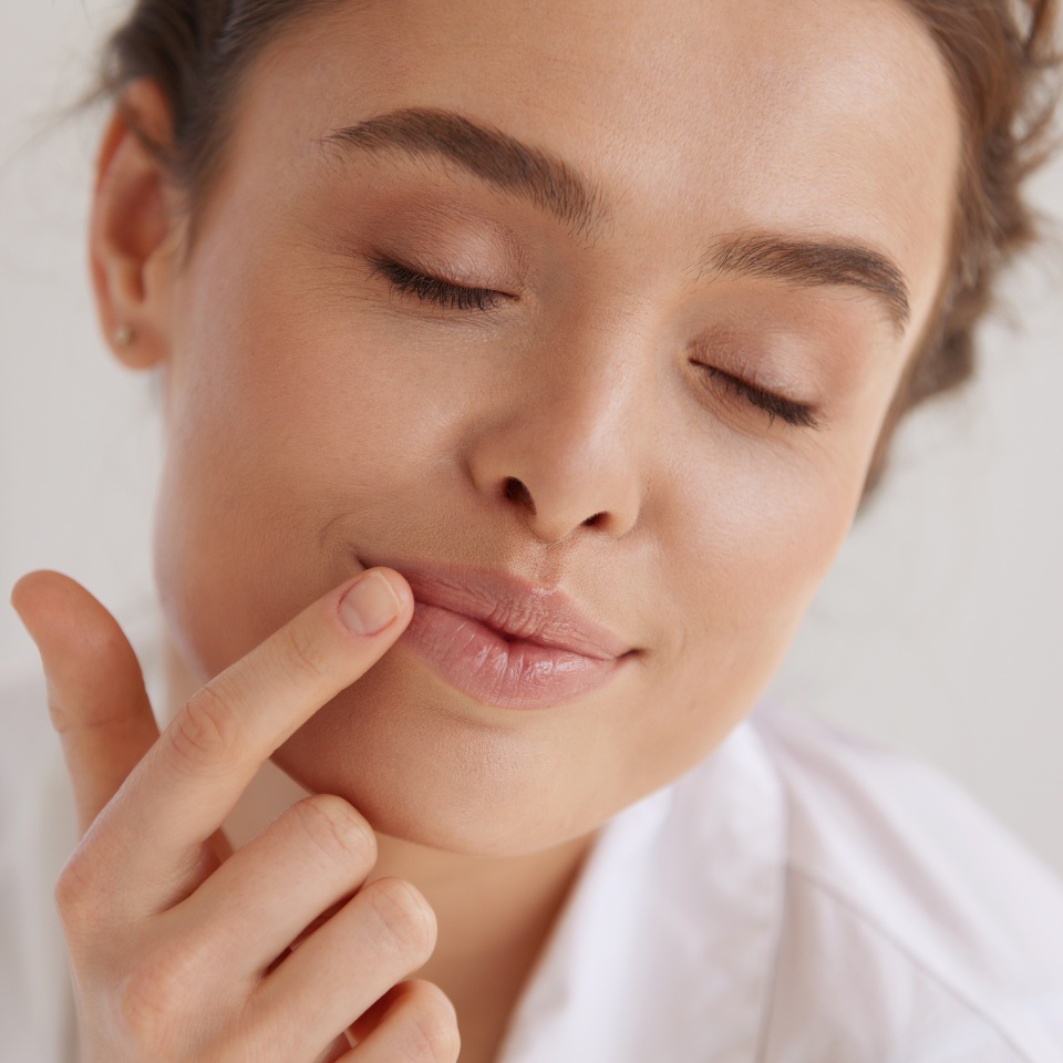Lips Protection. Beautiful Woman With Happy Face Applying Lip Balm. Portrait of Brunette Girl With Closed Eyes Using Hygienic Balm In Studio. Lips Care Concept