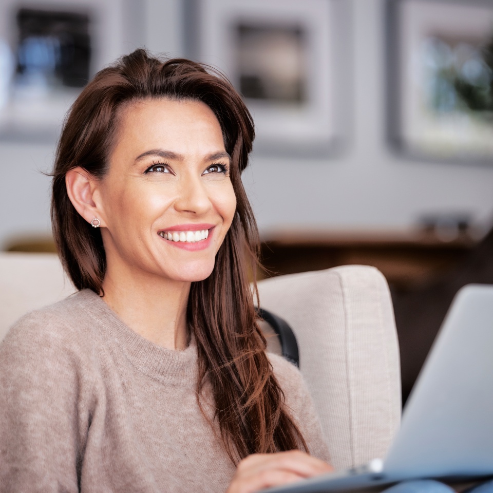 Close-up of an attractive middle aged woman sitting on armchair while using laptop. Beautiful female wearing casual clothes. Home office.