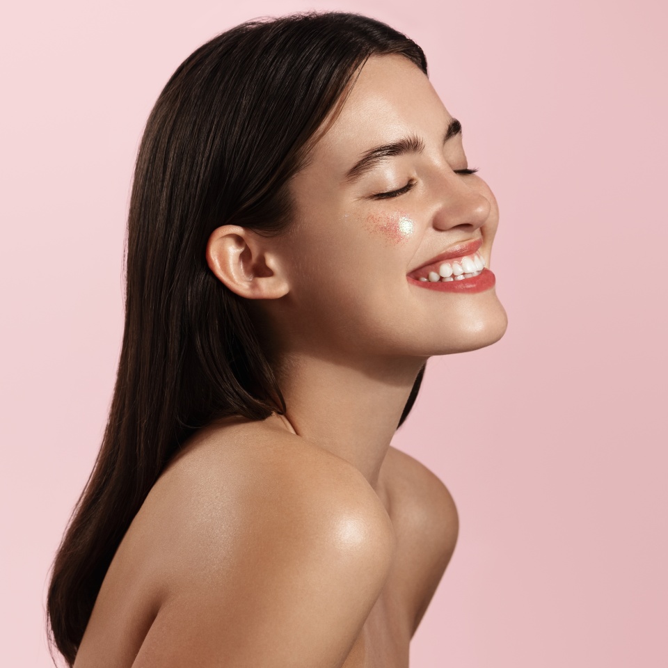 Haircare and beauty concept. Close up of happy smiling woman touches her natural hair, healthy glowing hair, white teeth and clean facial skin, standing against pink background.