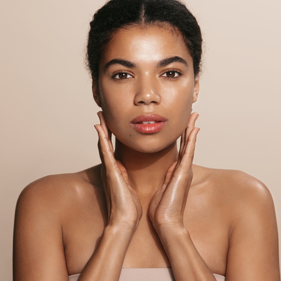 Close up shot of dark skinned woman with curly hair, appiles face cream on cheek, poses nude against brown background, cares of beauty, uses cosmetic product. Natural beauty concept.