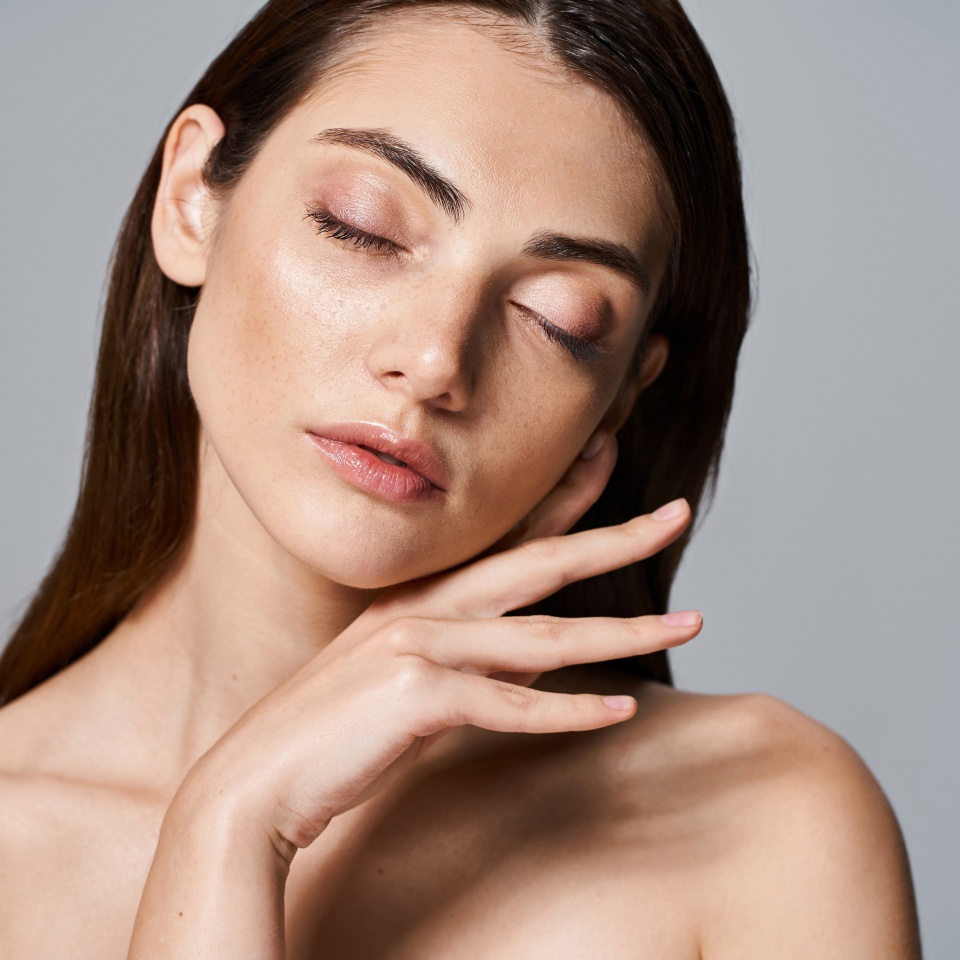A young, caucasian woman with brunette hair and clean skin posing nude with her eyes gently closed in a studio setting.
