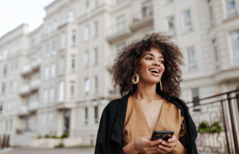 A woman with curly hair and large earrings stands outdoors in front of an apartment building, holding a smartphone and smiling.
