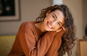 A person with curly hair, wearing a brown sweater, smiles while resting their head on their hands.