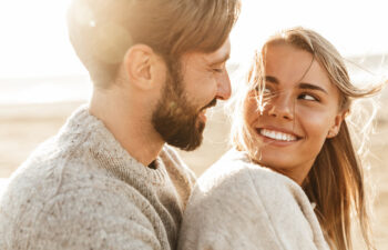 A man and woman wearing sweaters sit close together outdoors, smiling at each other in sunlight.