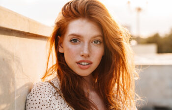 A woman with long red hair and freckles looks at the camera, standing outdoors near a concrete wall in soft sunlight.