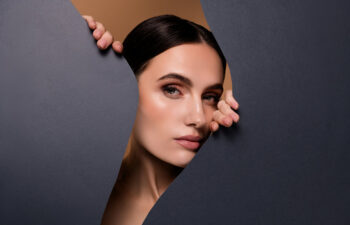 A woman with dark hair and natural makeup peers through a jagged opening in a dark paper backdrop.