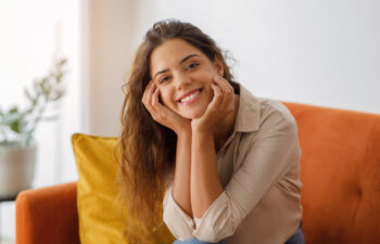A woman with long brown hair sits on an orange sofa, resting her chin on her hands and smiling at the camera. A plant is visible in the background.